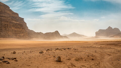 Durch eine weite Wüstenlandschaft weht Wind und wirbelt Sand auf.