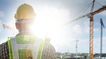 UV Belastung am Arbeitsplatz: ein Bauarbeiter steht mit dem Rücken zur Kamera und blickt über eine Baustelle. Dabei schaut er direkt in die strahlende Sonne. Bild von Adobe Stock