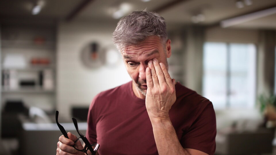 Ein Mann hat die Brille abgesetzt und reibt sich mit der linken Hand das Auge