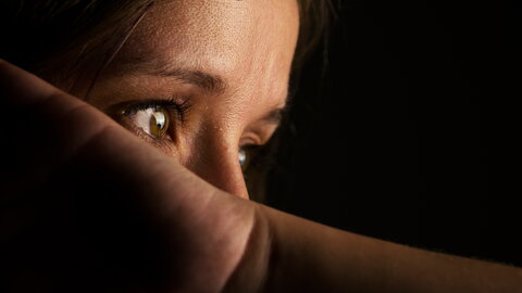 Nahaufnahme vom Gesicht einer Frau im Halbdunkel. Die Frau deckt den unteren Bereich ihres Gesichts mit dem Unterarm ab.